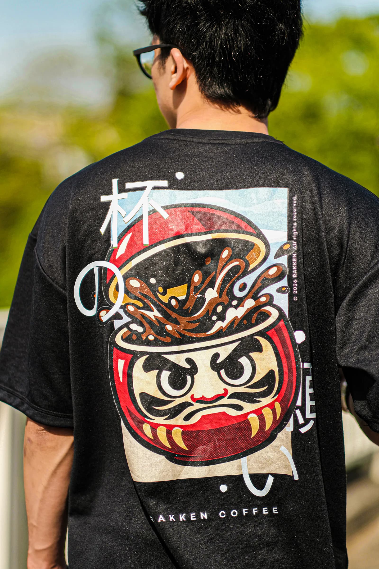 Kaos hitam RAKKEN dengan grafis Daruma dikenakan dari belakang
