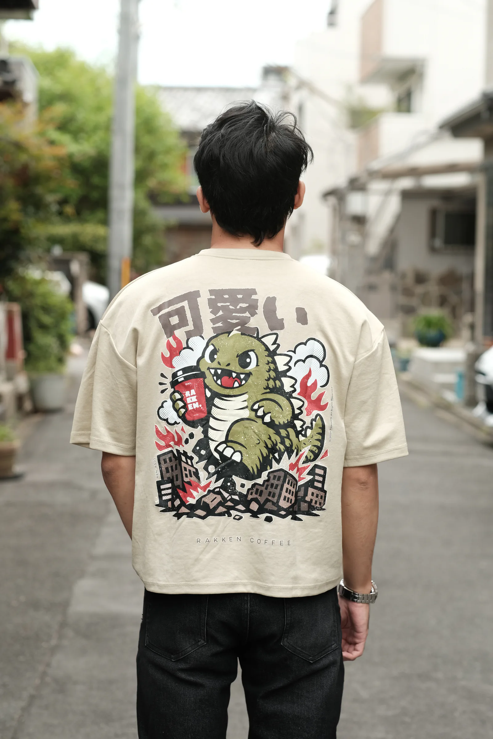 Kaos cream RAKKEN dengan grafis Godzilla dikenakan dari belakang